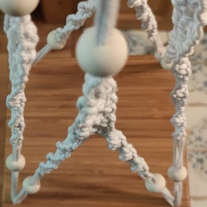 May include: Close-up of a macrame plant hanger with a light grey rope design. The hanger features wooden beads and a woven pattern. The background is a wooden surface.