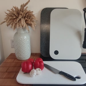 May include: A kitchen still life with three rectangular cutting boards in white and speckled grey. Red bell peppers, garlic, and a knife are arranged on a white board. A vase with wheat stalks is also present.