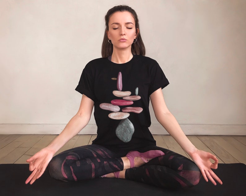 May include: A woman sits in a meditative pose wearing a black t-shirt with a graphic of a stack of colorful stones. She is wearing black leggings with a pink and black pattern.