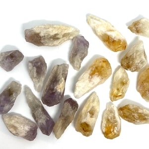 May include: A collection of raw, uncut crystals in shades of purple, yellow, and white. The crystals vary in size and shape, with a rough, natural texture. The arrangement is scattered on a white surface, showcasing the natural beauty of the gemstones.