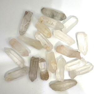 May include: A collection of raw quartz crystals in various shapes and sizes. The crystals range in color from clear to pale beige and light brown. Some crystals have a frosted appearance, while others are more transparent.