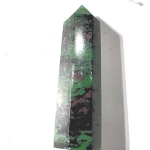 May include: A polished, rectangular gemstone obelisk. The stone features a mix of green, black, and red hues. The top is a pointed, pyramid shape. The obelisk is set against a white background.