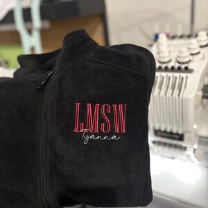 May include: Black fleece zip-up jacket with the embroidered text "LMSW Tyanna" in pink. The jacket has a high collar and a full-length zipper. The embroidery is centered on the left chest.