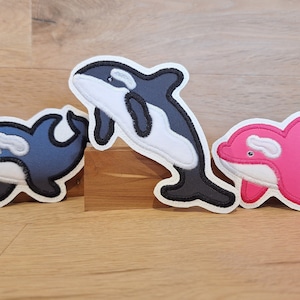 Könnte beinhalten: Drei Filzapplikationen von verschiedenen Delfinarten. Ein schwarz-weißer Orca, ein blauer Delfin und ein rosa Delfin.
