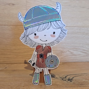 Könnte beinhalten: Eine weiße und graue gestickte Filzapplikation eines Comic-Charakters, der einen silbernen Helm mit Hörnern, eine rot-braune Tunika trägt und ein Schwert und einen Schild hält.