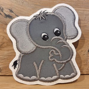 Könnte beinhalten: Eine gestickte Applikation eines grauen Elefanten mit weißem Umriss. Der Elefant hat einen eingerollten Rüssel und große Augen.