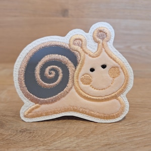Könnte beinhalten: Eine beige und braune gestickte Schnecken-Applikation auf weißem Hintergrund. Die Schnecke hat eine braune Schale mit einem Spiralmuster und einen beigefarbenen Körper mit einem lächelnden Gesicht.