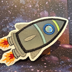 Könnte beinhalten: Ein grauer und weißer gestickter Raketen-Patch mit orangefarbenen Flammen. Die Rakete hat ein blaues und grünes Fenster mit einem holografischen Kreis in der Mitte.