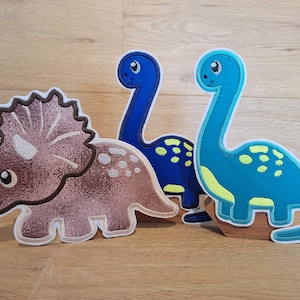 Könnte beinhalten: Drei Filz-Dinosaurierfiguren, ein braunes Triceratops, ein blauer Stegosaurus und ein türkisfarbener Brachiosaurus, stehen auf Holzsockeln.