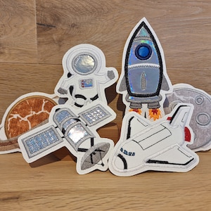Könnte beinhalten: Ein Set mit fünf Bügelbildern mit Weltraummotiven. Die Patches zeigen eine Rakete, einen Astronauten, ein Raumschiff, einen Satelliten und einen Planeten mit Ringen. Die Patches sind alle weiß mit silbernen und blauen Akzenten.
