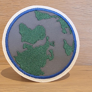 Könnte beinhalten: Ein kreisförmiger bestickter Aufnäher mit einer stilisierten Landkarte aus dunkelgrünem Garn auf grauem Hintergrund. Das Design ist von einem leuchtend blauen kreisförmigen Rand umschlossen und mit einer cremefarbenen Außenkante versehen.
