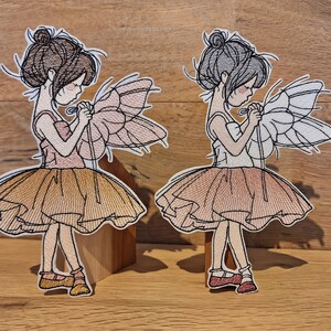 Könnte beinhalten: Zwei gestickte Aufnäher von Ballerinas mit Flügeln. Eine Ballerina trägt einen rosa-braunen Tutu und hat braunes Haar. Die andere Ballerina trägt einen weiß-braunen Tutu und hat graues Haar.
