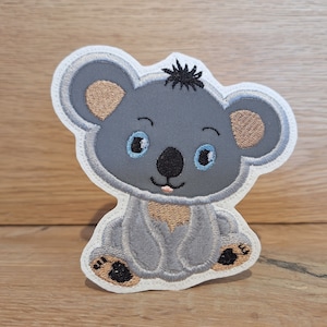 Könnte beinhalten: Ein grauer Koalabär mit braunen Ohren und blauen Augen sitzt auf einem weißen Hintergrund. Der Koala ist mit schwarzen Nähten gestickt.