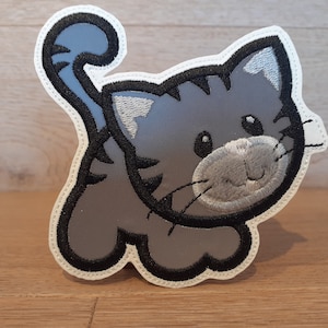Könnte beinhalten: Eine graue gestickte Katze mit schwarzen Umrissen und weißem Hintergrund. Die Katze hat ein süßes, cartoonartiges Gesicht mit großen Augen und einem kleinen Lächeln.