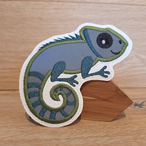Könnte beinhalten: Gestickter Aufnäher eines grauen Chamäleons mit teal- und grünen Akzenten. Das Chamäleon hat ein lächelndes Gesicht und einen eingerollten Schwanz. Der Aufnäher befindet sich auf weißem Hintergrund und ist auf einem Holzständer befestigt.