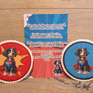 Könnte beinhalten: Zwei runde, rote und blaue Filzuntersetzer mit weißem Rand. Jeder Untersetzer zeigt einen Comic-Hund, der ein Superhelden-Kostüm mit einem roten Umhang trägt. Der Text auf der Karte lautet: "Ich schenk dir einen Superheld, der dir stets die Treue hält. Ein Abenteuer jeden Tag, ist was er am liebsten mag. Und an nicht ganz so guten Tagen, kann er deine Sorgen tragen. d denkst du mal, du schaff was nicht, in glaubt dein Superheld dich."