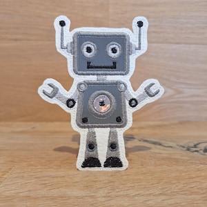 Könnte beinhalten: Ein grauer Roboter vor weißem Hintergrund. Der Roboter hat einen runden Kopf mit zwei schwarzen Augen, einem schwarzen Mund und einem silbernen Körper. Der Roboter hat zwei Arme mit silbernen Händen und zwei Beine mit schwarzen Füßen.