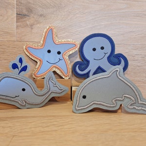 Könnte beinhalten: Vier Filzapplikationen von Meeresbewohnern: ein blauer Oktopus, ein grauer Wal, ein grauer Delfin und ein goldener Seestern. Jede Applikation hat ein lächelndes Gesicht.
