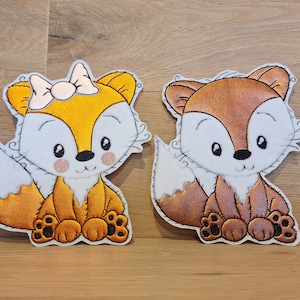 Könnte beinhalten: Zwei gestickte Fuchsflicken, einer mit gelbem und orangefarbenem Körper und einer weißen Schleife, der andere mit braunem Körper. Beide Füchse sitzen mit ihren Schwänzen hinter sich eingerollt.
