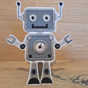 Könnte beinhalten: Gestickter Aufnäher eines freundlichen Roboters in Grau-, Weiß- und Schwarztönen. Der Roboter hat einen quadratischen Kopf mit zwei Antennen, kreisförmigen Augen und schraubenförmigen Armen. Ein kreisförmiges Detail auf der Brust hat einen holografischen Effekt.