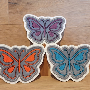Könnte beinhalten: Drei gestickte Bügelbilder in Form von Schmetterlingen. Die Schmetterlinge sind grau umrandet und mit verschiedenen Farben gefüllt: lila, orange und blau.