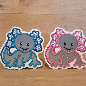 Könnte beinhalten: Zwei gestickte Axolotl-Aufnäher. Einer ist grau mit blauen und violetten Akzenten, der andere grau mit rosa Akzenten. Beide haben lächelnde Gesichter und sind auf weißem Grund.