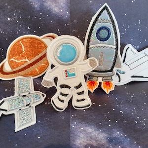 Könnte beinhalten: Fünf gestickte Bügelbilder mit Weltraummotiven. Die Patches zeigen eine Rakete, ein Raumschiff, einen Astronauten, einen Planeten mit Ringen und einen Satelliten. Die Patches sind alle weiß mit bunten Details.