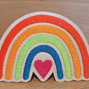 Könnte beinhalten: Ein gestickter Patch in Regenbogenfarben mit einem pinkfarbenen Herzen in der Mitte. Der Regenbogen hat sechs Farben: Rot, Orange, Gelb, Grün, Blau und Lila.