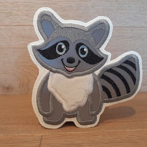 Könnte beinhalten: Ein gestickter Waschbär-Applikateur in Grau und Weiß mit einem schwarz-weiß gestreiften Schwanz. Der Waschbär hat einen weißen Bauch und schwarze Augen.