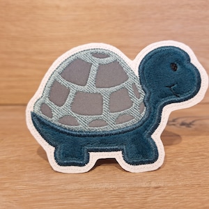 Könnte beinhalten: Ein gestickter Schildpatt-Applikations-Patch in Blau und Grau. Die Schildkröte hat einen Panzer mit einem Muster aus Quadraten und ein lächelndes Gesicht.