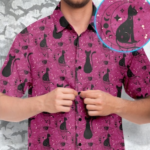 May include: A pink button-up shirt with a black cat and moon pattern. The shirt has a short sleeve and a button-up collar.