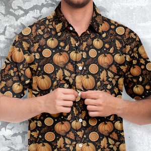 Puede incluir: Una camisa negra de manga corta con botones y un estampado de temática otoñal. El estampado presenta calabazas naranjas, hojas marrones y rodajas de naranja sobre un fondo negro.