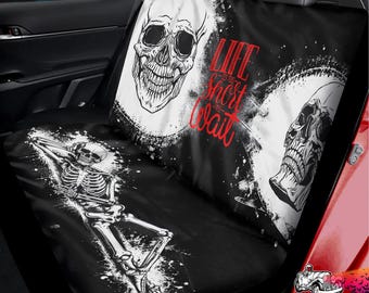 May include: A black and white car seat cover featuring a graphic print of two skulls and a reclining skeleton. The text "LIFE IS TOO SHORT TO WAIT" is printed in red script in the center. The design includes white splatter accents on a black background.