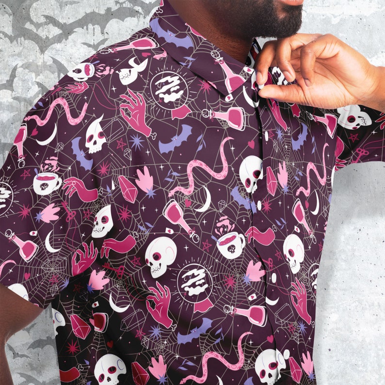 Puede incluir: Una camisa abotonada de color p&uacute;rpura oscuro con un patr&oacute;n repetido de calaveras blancas, manos rosas, botellas de pociones, murci&eacute;lagos y telara&ntilde;as. La camisa tiene cuello y mangas cortas. El dise&ntilde;o incluye acentos rosas, blancos y morados claros.