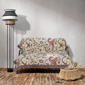 May include: A cream-colored throw blanket with a woven design of red dragons and floral vines. The blanket is draped over a wooden couch with a black metal frame. A black and white lamp with a wicker shade stands next to the couch. A woven basket sits on the floor in front of the couch.
