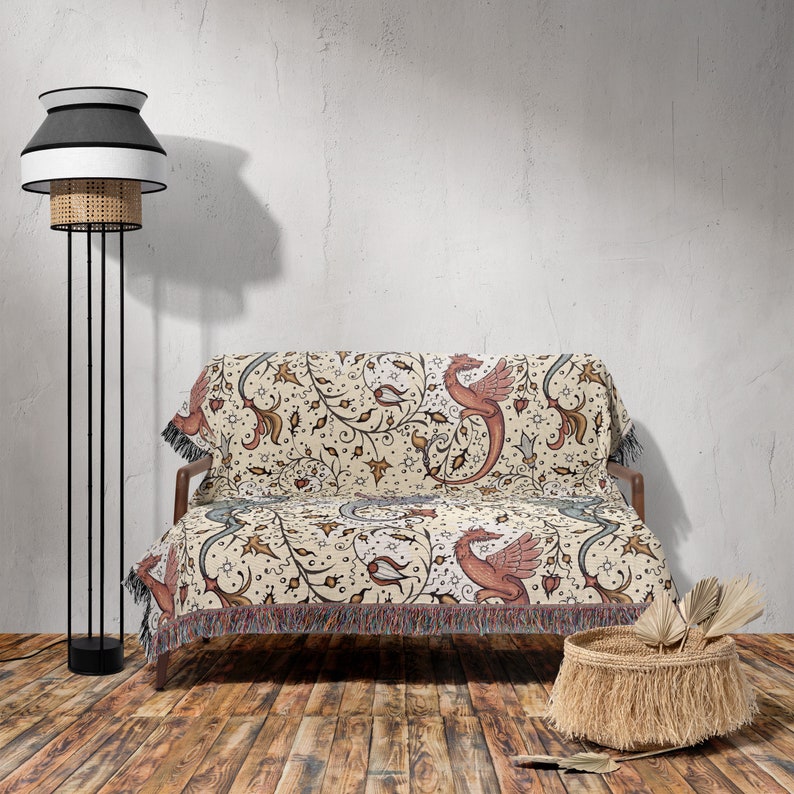 May include: A cream-colored throw blanket with a woven design of red dragons and floral vines. The blanket is draped over a wooden couch with a black metal frame. A black and white lamp with a wicker shade stands next to the couch. A woven basket sits on the floor in front of the couch.