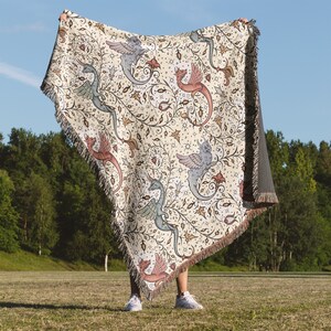 May include: A cream-colored throw blanket with a tapestry design featuring stylized dragons and floral patterns. The blanket has a fringe border and is being held up by a person.