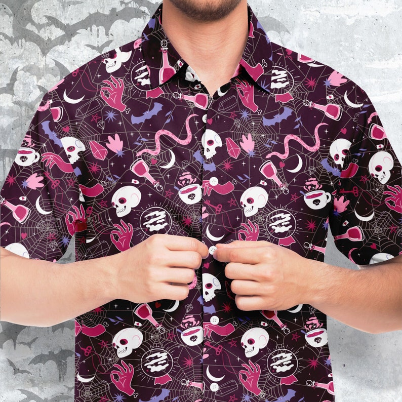 Puede incluir: Camisa abotonada de color p&uacute;rpura oscuro con un patr&oacute;n repetido de calaveras blancas, manos rosas, murci&eacute;lagos y telara&ntilde;as. La camisa tiene un cuello cl&aacute;sico y mangas cortas. El dise&ntilde;o incluye acentos rosas y morados.