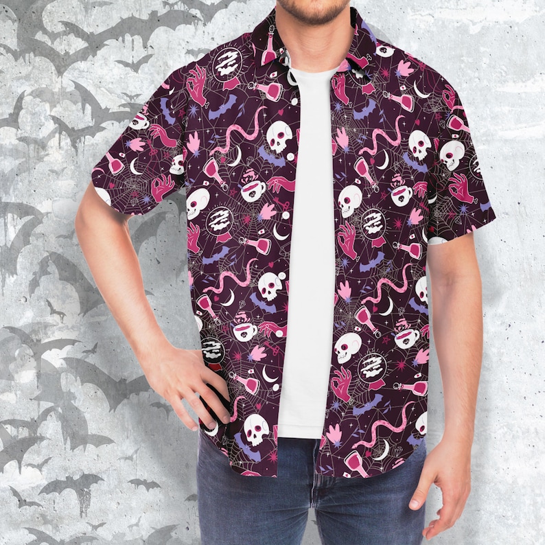 Puede incluir: Camisa de manga corta con fondo morado oscuro y un patr&oacute;n repetido de calaveras blancas, botellas de pociones, manos, murci&eacute;lagos y telara&ntilde;as. La camisa est&aacute; abierta, revelando una camiseta blanca.