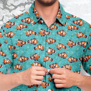Può includere: Una camicia a bottoni blu turchese con un motivo ripetitivo di pesci pagliaccio arancioni e bianchi. La camicia ha una chiusura a bottoni e maniche corte.