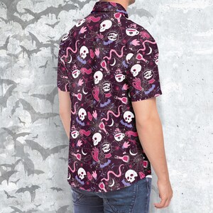 Puede incluir: Camisa de manga corta con fondo morado oscuro y un patr&oacute;n repetido de calaveras blancas, manos rosas, botellas de pociones y telara&ntilde;as. La camisa est&aacute; hecha de tela ligera, adecuada para ropa casual.