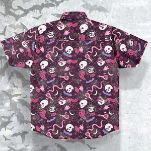 Puede incluir: Camisa de manga corta con botones, con un fondo morado oscuro y un patr&oacute;n repetido de calaveras blancas, botellas de pociones, manos y telara&ntilde;as en rosa, blanco y morado. La camisa tiene un cuello cl&aacute;sico y dos bolsillos en el pecho.
