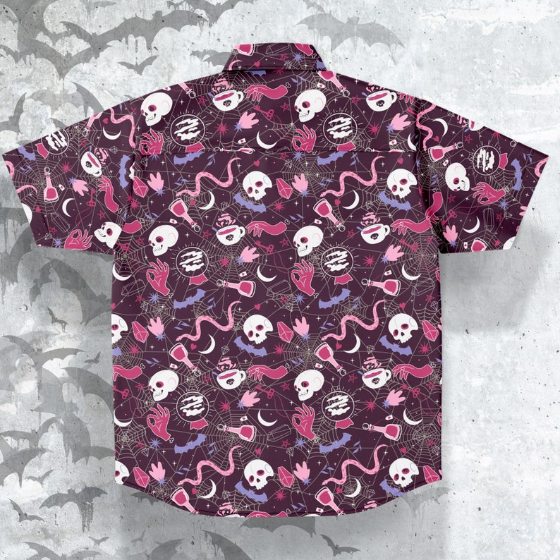 Puede incluir: Camisa de manga corta con botones, con un fondo morado oscuro y un patr&oacute;n repetido de calaveras blancas, botellas de pociones, manos y telara&ntilde;as en rosa, blanco y morado. La camisa tiene un cuello cl&aacute;sico y dos bolsillos en el pecho.