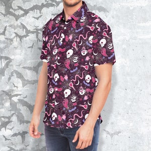 Puede incluir: Camisa de manga corta con un patr&oacute;n morado oscuro con calaveras blancas, manos rosas, murci&eacute;lagos, botellas de pociones y telara&ntilde;as. La camisa tiene un cuello cl&aacute;sico y se usa con vaqueros azules.