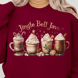 Könnte beinhalten: Ein weinrotes Sweatshirt mit dem Text "Jingle Bell Java" in goldener Schrift. Die Grafik zeigt vier Tassen festlicher Kaffeegetränke mit Schlagsahne, Zuckerstangen und Lebkuchen. Ein gemütliches, saisonales Design.