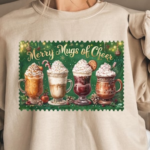 Peut inclure: Un sweat-shirt beige avec un motif festif. Le motif représente quatre tasses de boissons chaudes garnies de crème fouettée, d'un sucre d'orge et d'un biscuit. Le texte "Merry Mugs of Cheer" est au-dessus des tasses sur un fond vert.