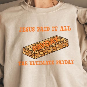 May include: Beige sweatshirt with orange text that reads "JESUS PAID IT ALL" above a graphic of a candy bar with the word "PAYDAY" on it. Below the candy bar is the text "THE ULTIMATE PAYDAY".