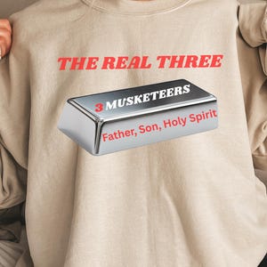 May include: Beige sweatshirt with the text "THE REAL THREE" in red. A silver bar graphic reads "3 MUSKETEERS" and "Father, Son, Holy Spirit" in red. The sweatshirt is a neutral color.