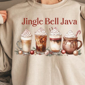 Peut inclure: Sweat-shirt beige avec l'inscription rouge "Jingle Bell Java". Le motif représente quatre boissons au café avec de la crème fouettée, des décorations de Noël et de la verdure. Un des mugs contient un sucre d'orge.