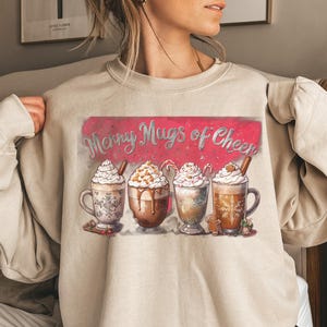 Peut inclure: Sweat-shirt beige avec un motif festif. Le motif présente quatre tasses de boissons chaudes, chacune garnie de crème fouettée et de décorations festives. Le texte "Merry Mugs of Cheer" est affiché sur un fond rouge.